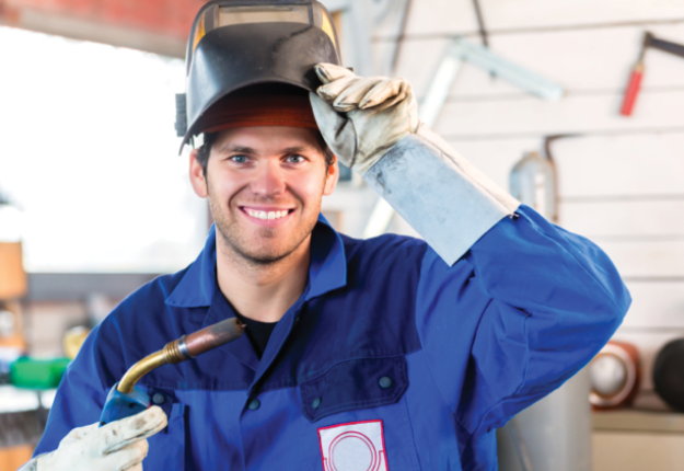 A man with a welding helmet and gloves, smiling as he works on a project.
