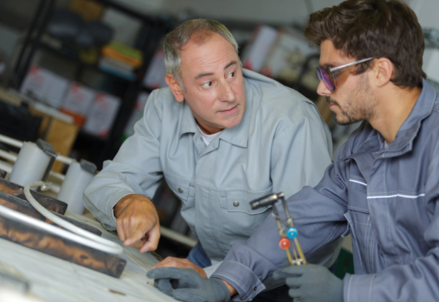 5 Two men in a factory, one a mentor and one a student, working on a metal object, using tools and machinery to complete their task efficiently.