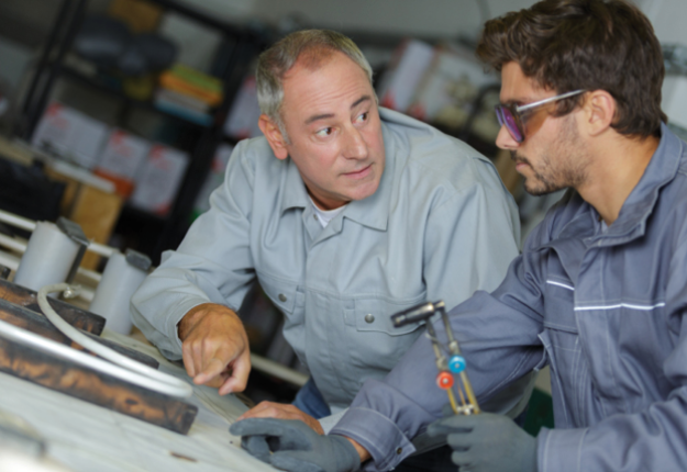 Two men in a factory, one a mentor and one a student, working on a metal object, using tools and machinery to complete their task efficiently.