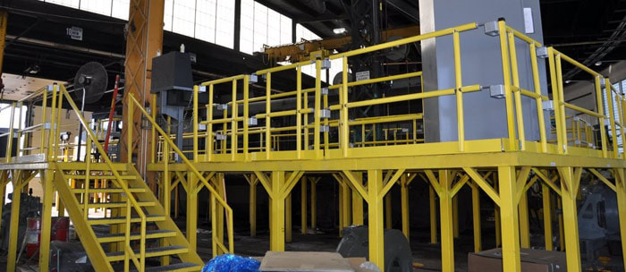 The inside of a industrial complex with yellow aluminum stairs that lead to an elevated platform with yellow handrails.