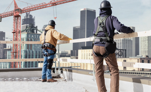 Two construction workers on a roof, observing a construction site with the city in the background while leaning on a portable guardrail system.